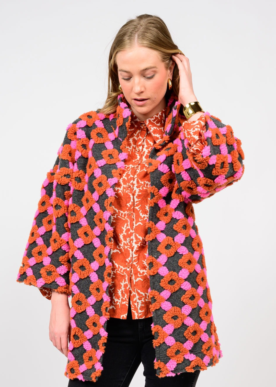 Woman wearing a pink, grey, and organe coat over an orange top and black pants in front of a white background