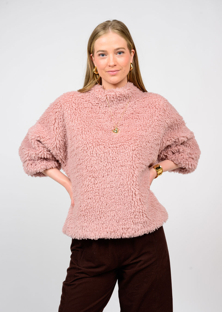 Woman with hands on hips wearing a pink sherpa top with 3/4 length sleeves and black jeans in front of a white background