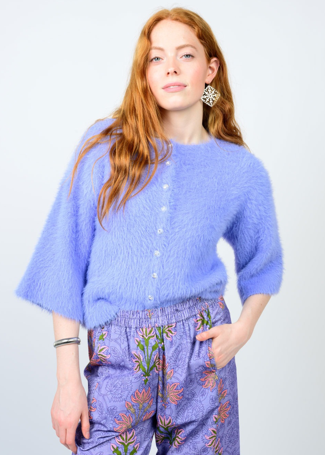 Woman wearing a light blue fuzzy cardigan with 3/4 length sleeves and purple floral pants against a white background
