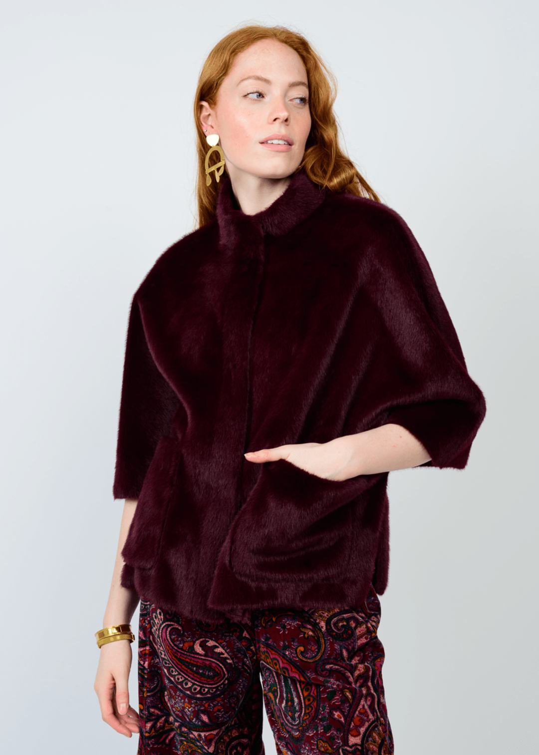 Woman wearing a deep wine colored fur jacket with 3/4 length sleeves and wine colored floral pants against a white background
