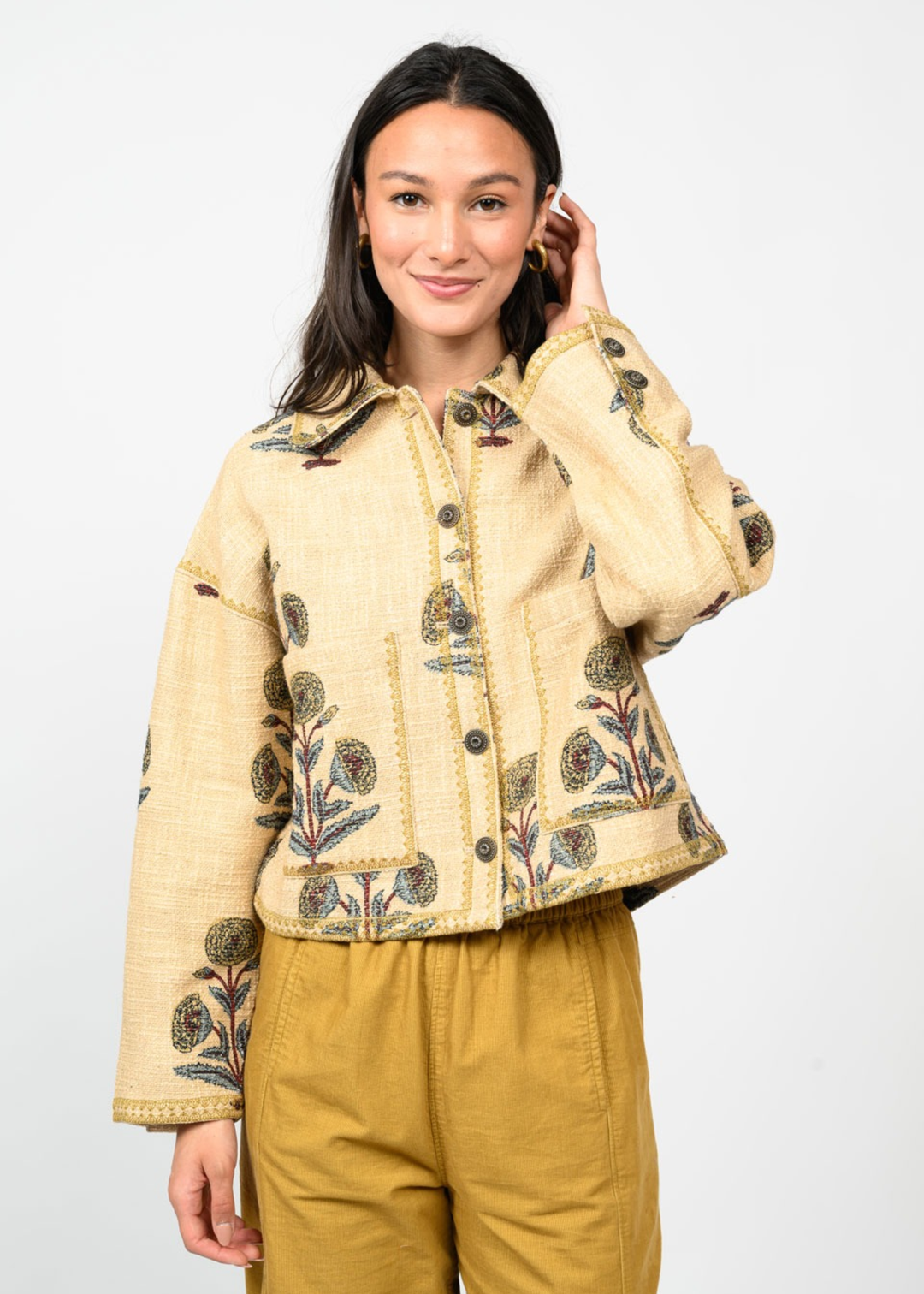Woman wearing a tan jacket with a floral print and gold pants in front of a white background