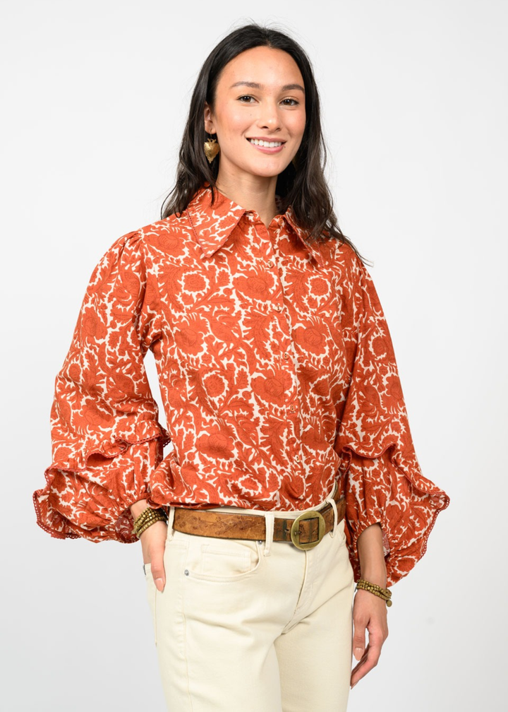 Woman wearing an orange floral patterned button down top with hand in pocket in front of a white background