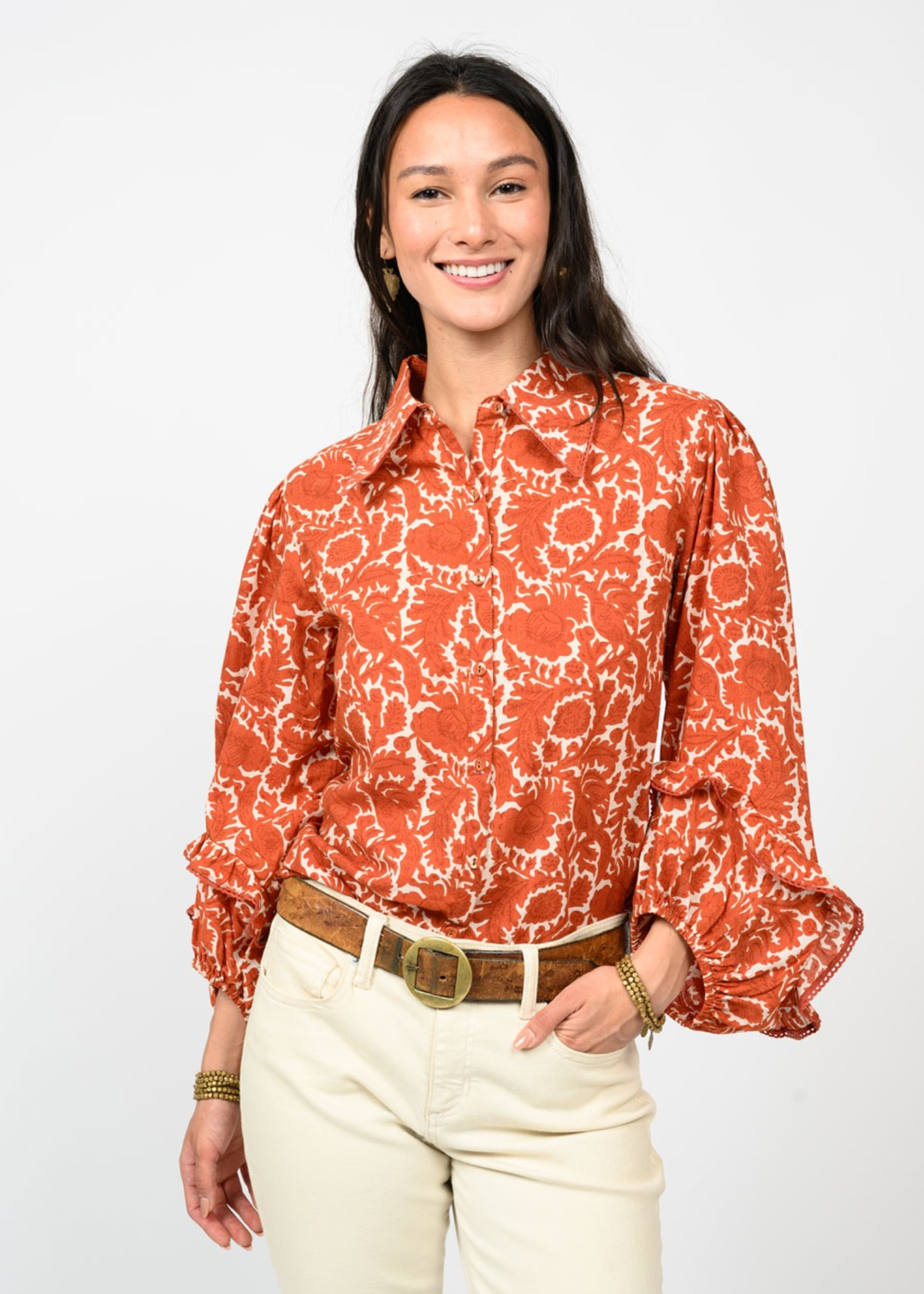 Woman wearing an orange floral patterned button down top with hand in pocket in front of a white background