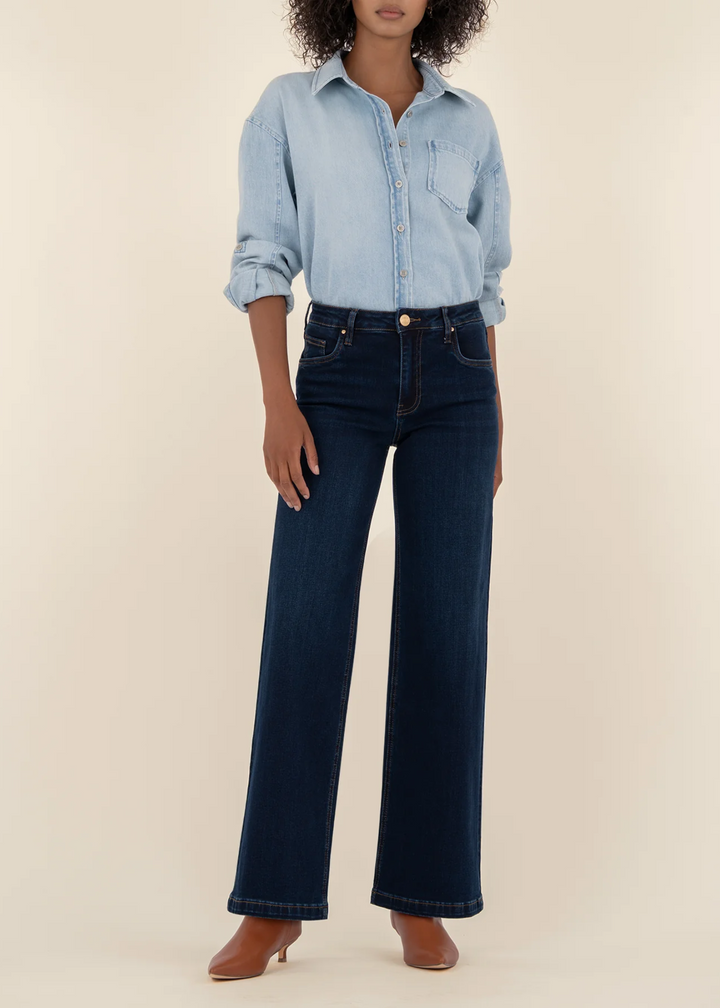 Woman wearing a light blue denim button down with dark blue wide leg jeans standing in front of a tan background