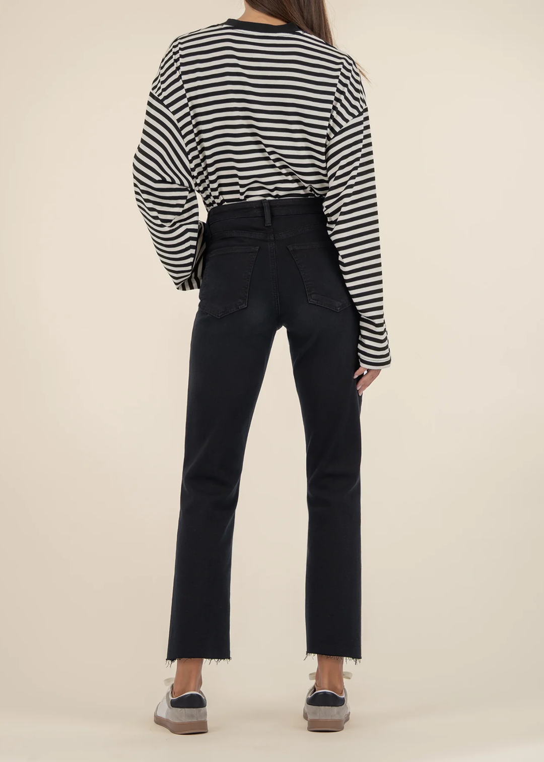 Woman turned around wearing black jeans with a black and white striped sweater and white sneakers standing in front of a white background