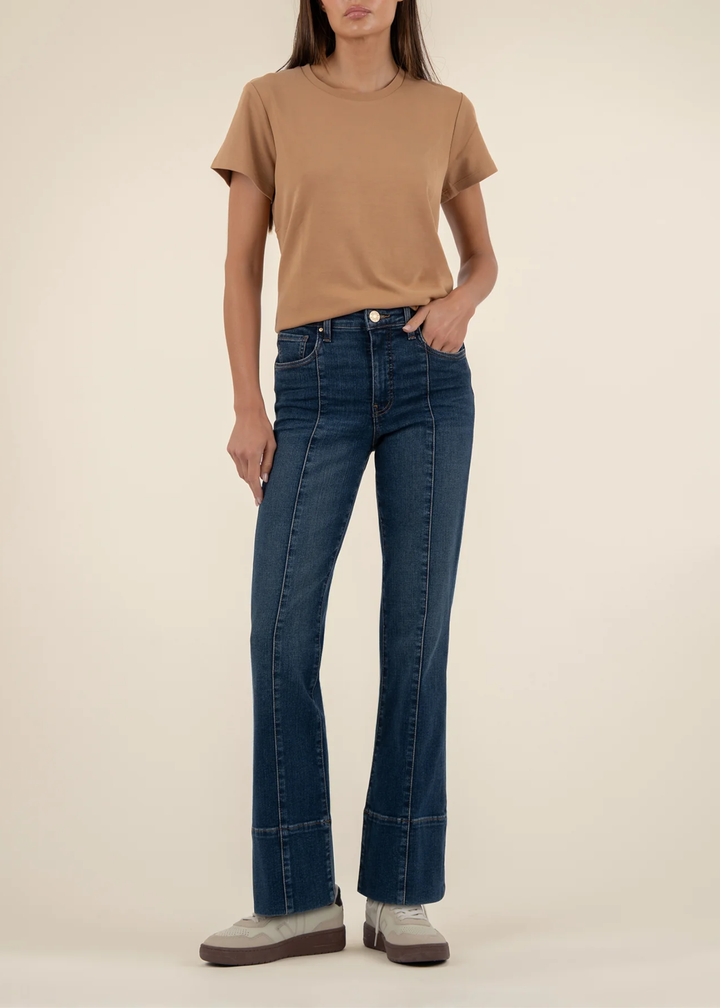 Woman wearing dark wash jeans with a front seam and a tan t-shirt with white shoes standing in front of a white background