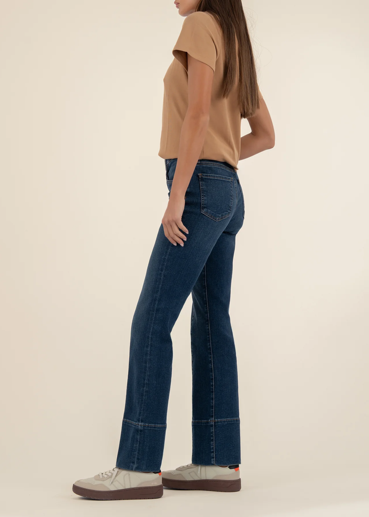 Woman turned to the side wearing dark wash jeans with a front seam and a tan t-shirt with white shoes standing in front of a white background