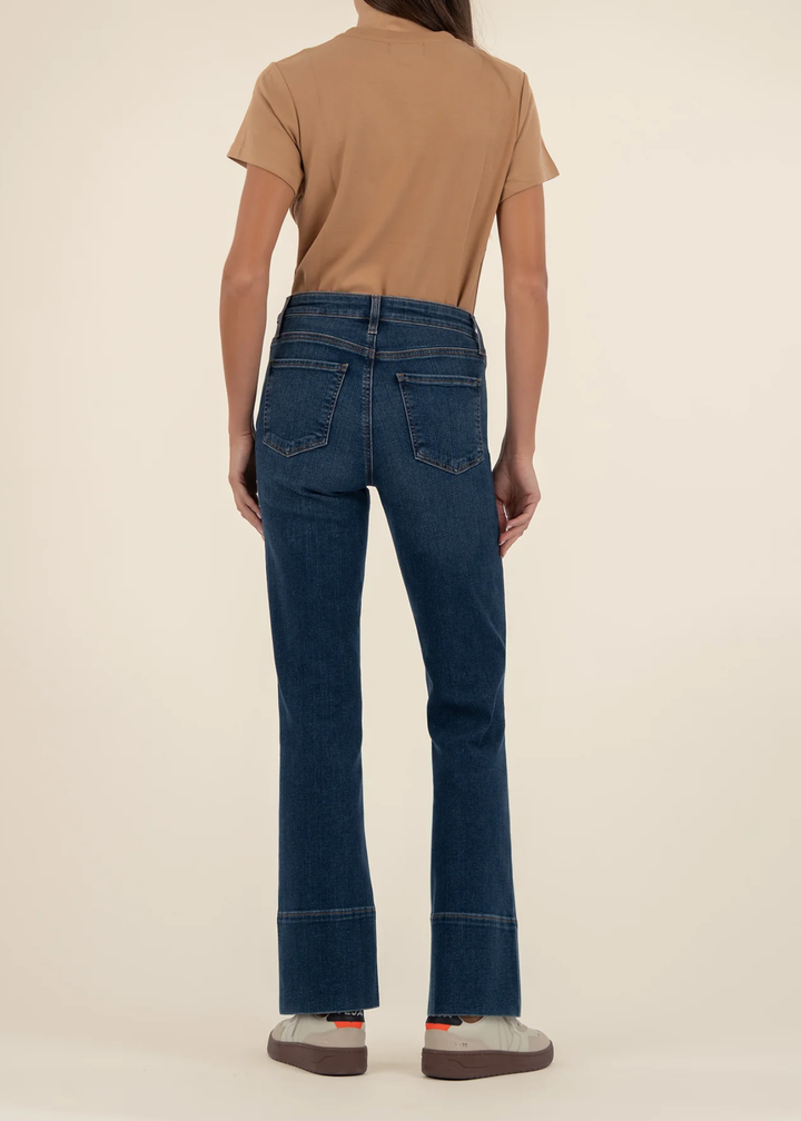 Woman turned around wearing dark wash jeans with a front seam and a tan t-shirt with white shoes standing in front of a white background