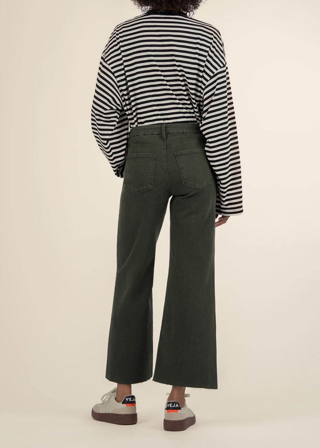 Woman turned around wearing dark green jeans with a black and white striped sweater and white sneakers in front of a white background