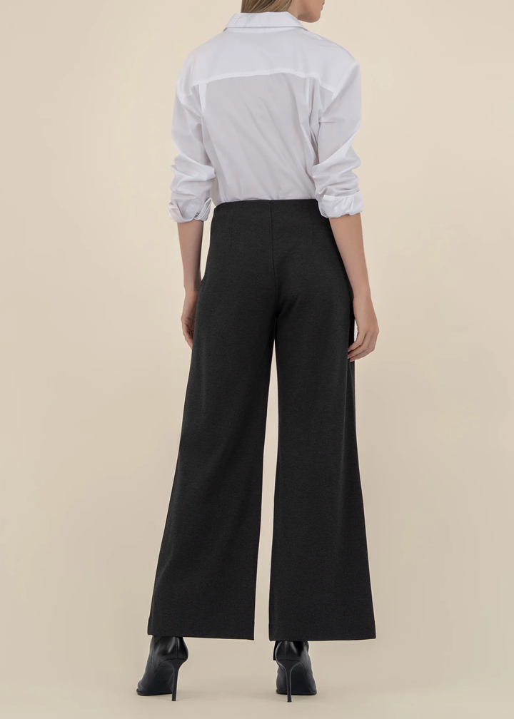 Woman turned around wearing a white button down with charcoal grey wide leg pants and black heeled boots in front of a tan background