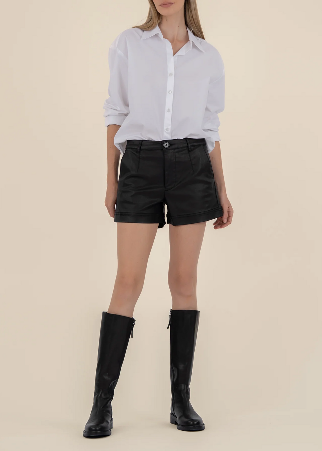 Woman wearing a white button down top with black shorts and tall black boots standing in front of a tan background