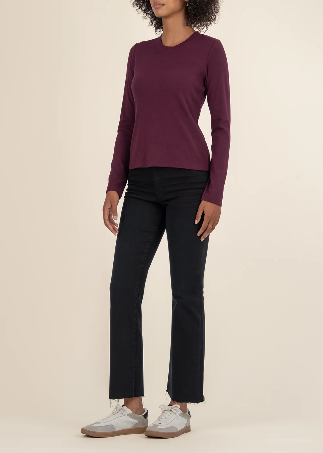 Woman turned to the side wearing a maroon long sleeve tee with black jeans and white sneakers in front of a white background