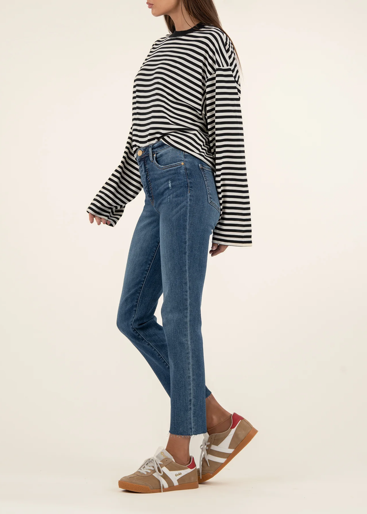 Woman turned to the side wearing medium wash blue ankle straight jeans with tan sneakers and a black and white striped sweater standing in front of a white background