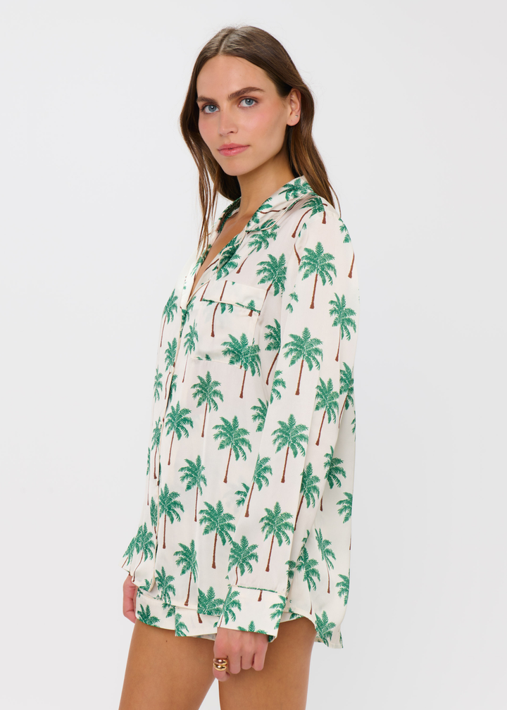 Woman turned to the side wearing a palm tree patterned pajama set with shorts and a long sleeve top against a white background
