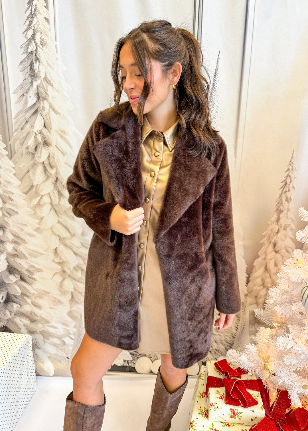 Woman wearing a brown faux fur coat in a festive setting with Christmas trees and presents.