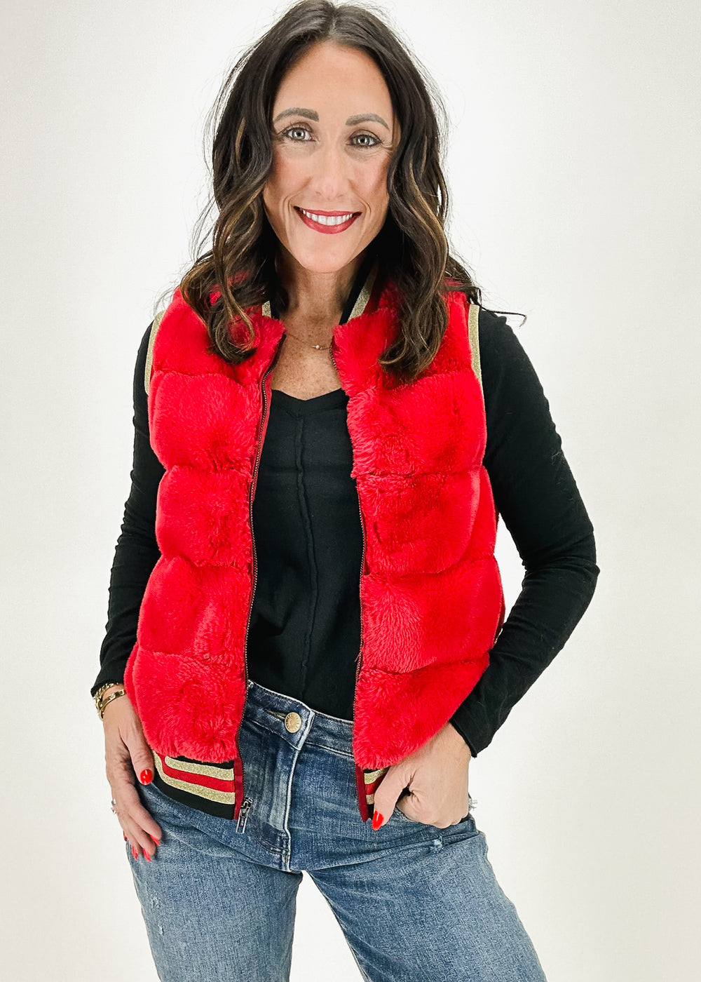 A woman wearing a red faux fur vest with gold trim and zipper closure, styled with a black top and jeans for a bold and cozy look.