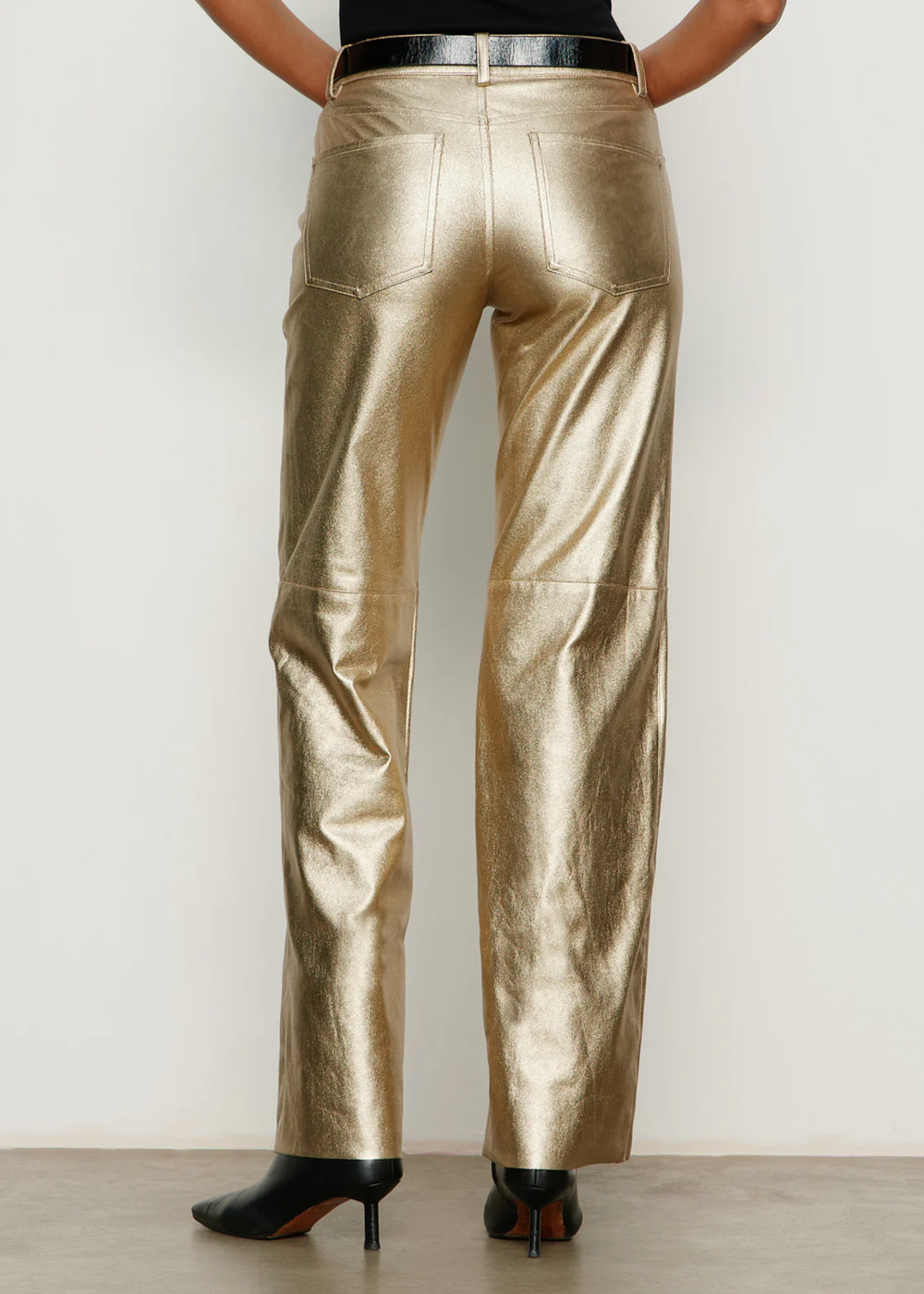 Woman turned around wearing gold pants with black boots and a black top against a white wall