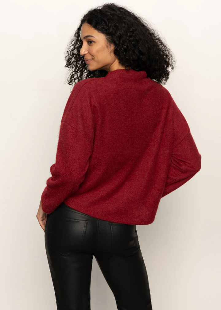 Woman turned around wearing a crimson red sweater with black pants standing in front of a white background