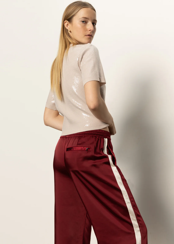 Woman turned to the side wearing red satin pants with a white side stripe and a tan sequined top