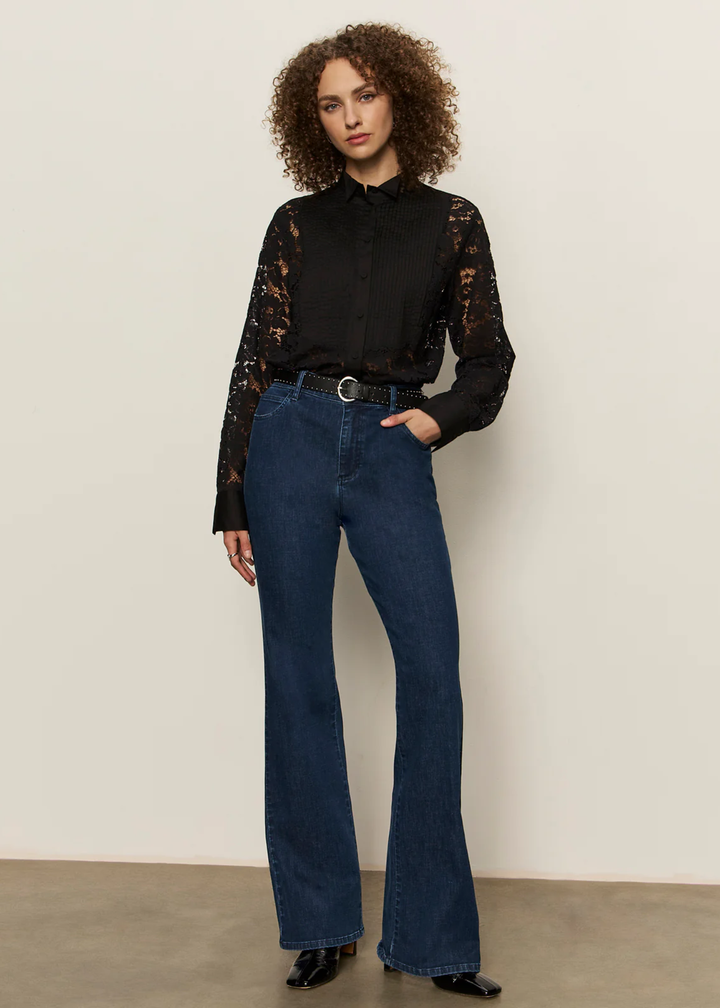 Woman with hand in pocket wearing a black button down shirt with lace sleeves and back with jeans against a white background