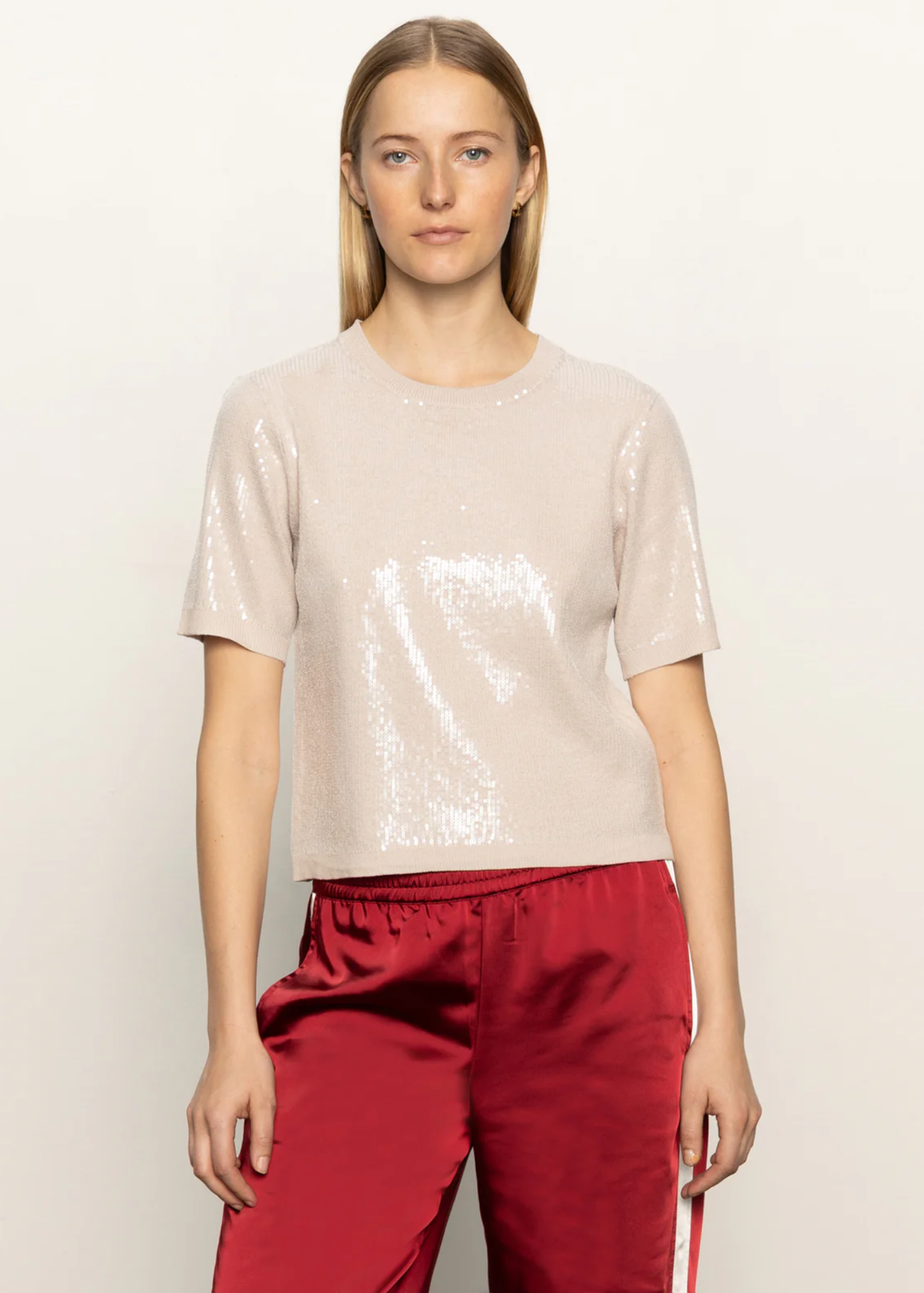 Woman wearing a cream sequined short sleeve top with red satin pants standing in front of a white background