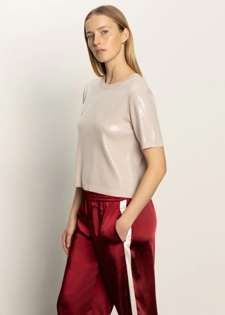 Woman turned to the side wearing a cream sequined short sleeve top with red satin pants standing in front of a white background