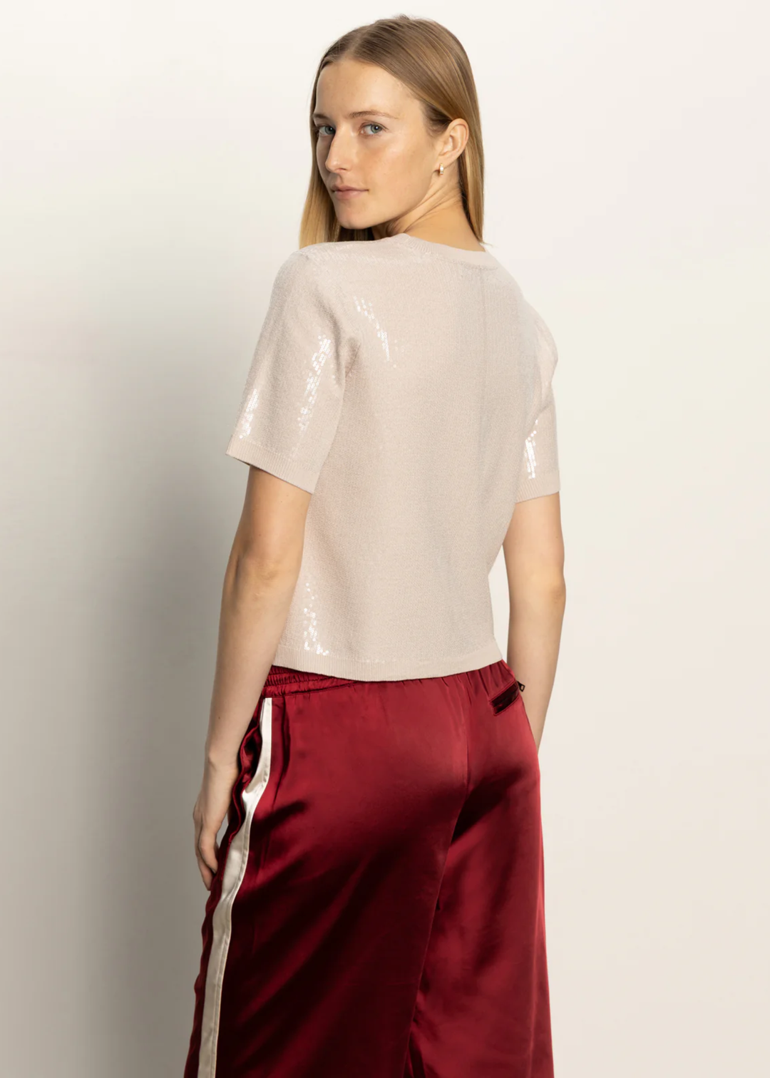 Woman turned around wearing a cream sequined short sleeve top with red satin pants standing in front of a white background