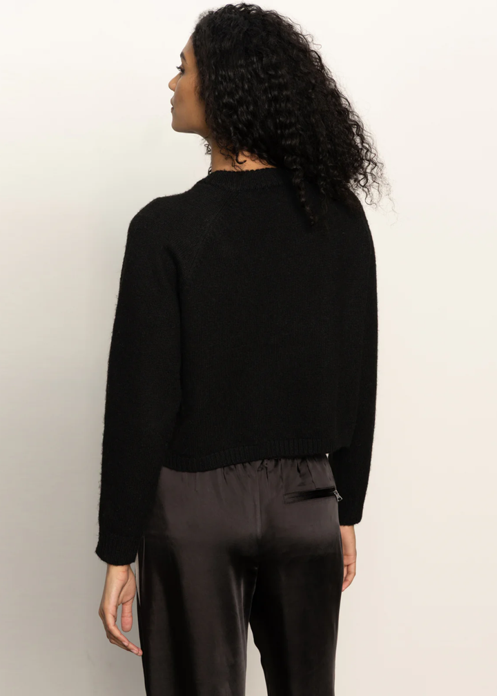 Woman turned around wearing a black sweater and black pants standing in front of a white background