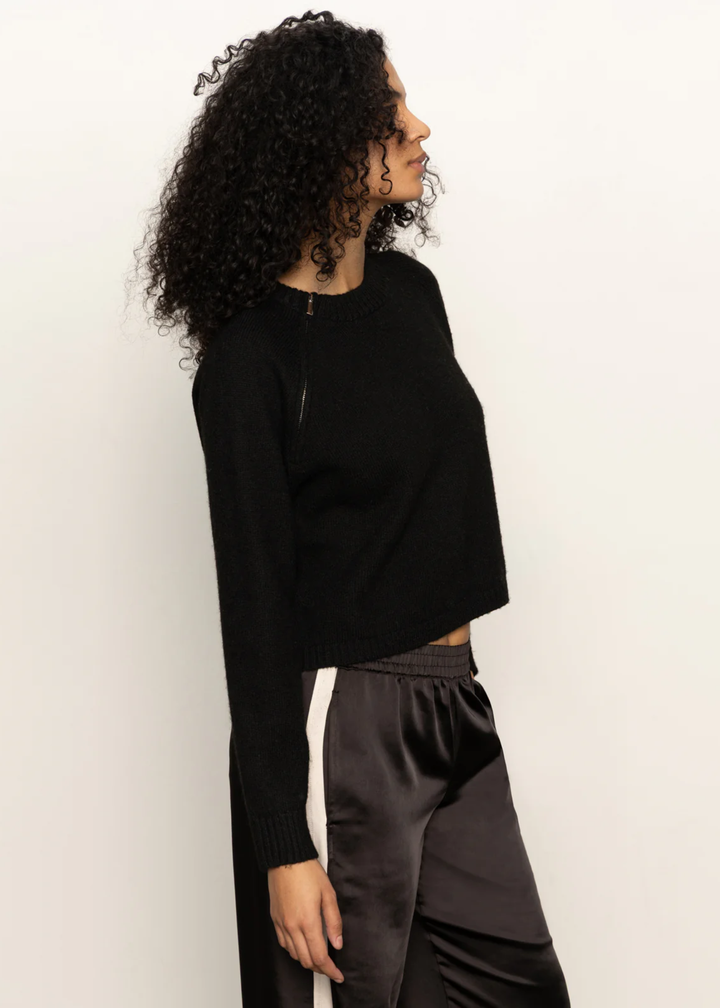 Woman turned to the side wearing a black sweater and black pants with a white side stripe standing in front of a white background
