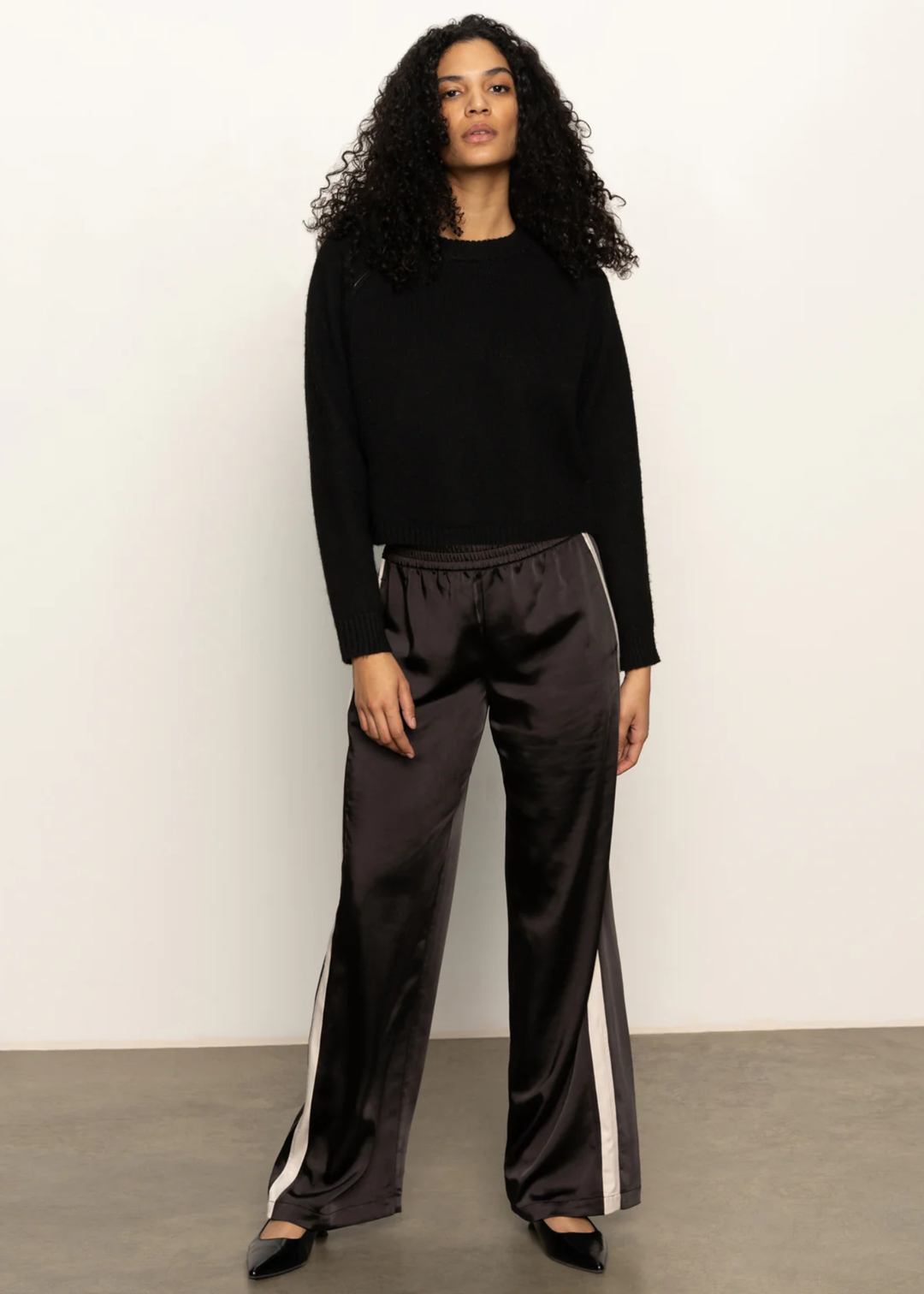 Woman wearing a black sweater and black pants with white side stripes and black flats standing in front of a white background