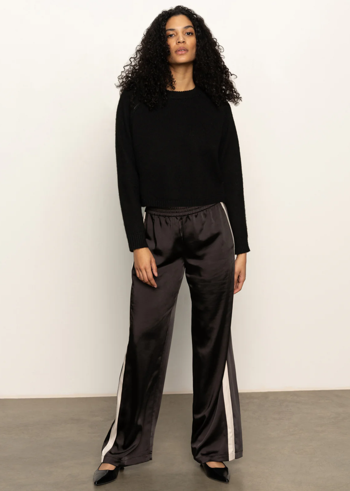 Woman wearing a black sweater and black pants with white side stripes and black flats standing in front of a white background