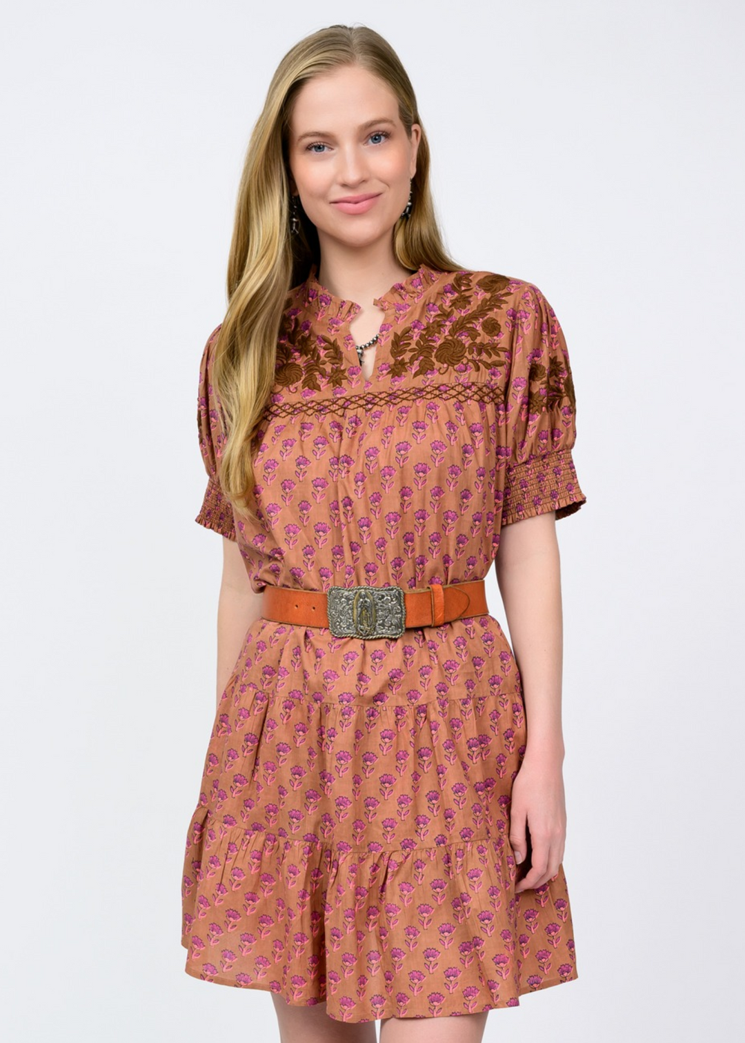 Woman wearing a light brown dress with a pink floral pattern, a v-neck, and brown embroidery in front of a white background