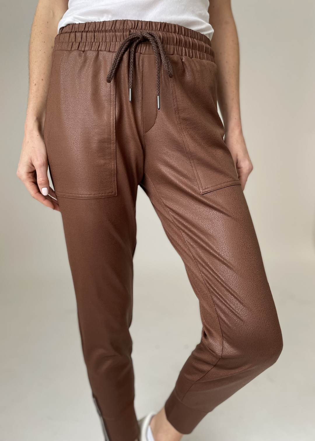 Woman wearing brown joggers with a white shirt in front of a beige background