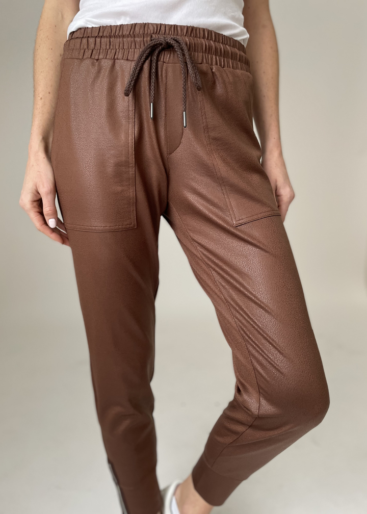 Woman wearing brown joggers with a white shirt in front of a beige background