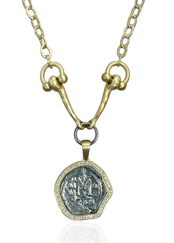 A gold horsebit chain necklace with a silver coin pendant encase by CZs on a white background