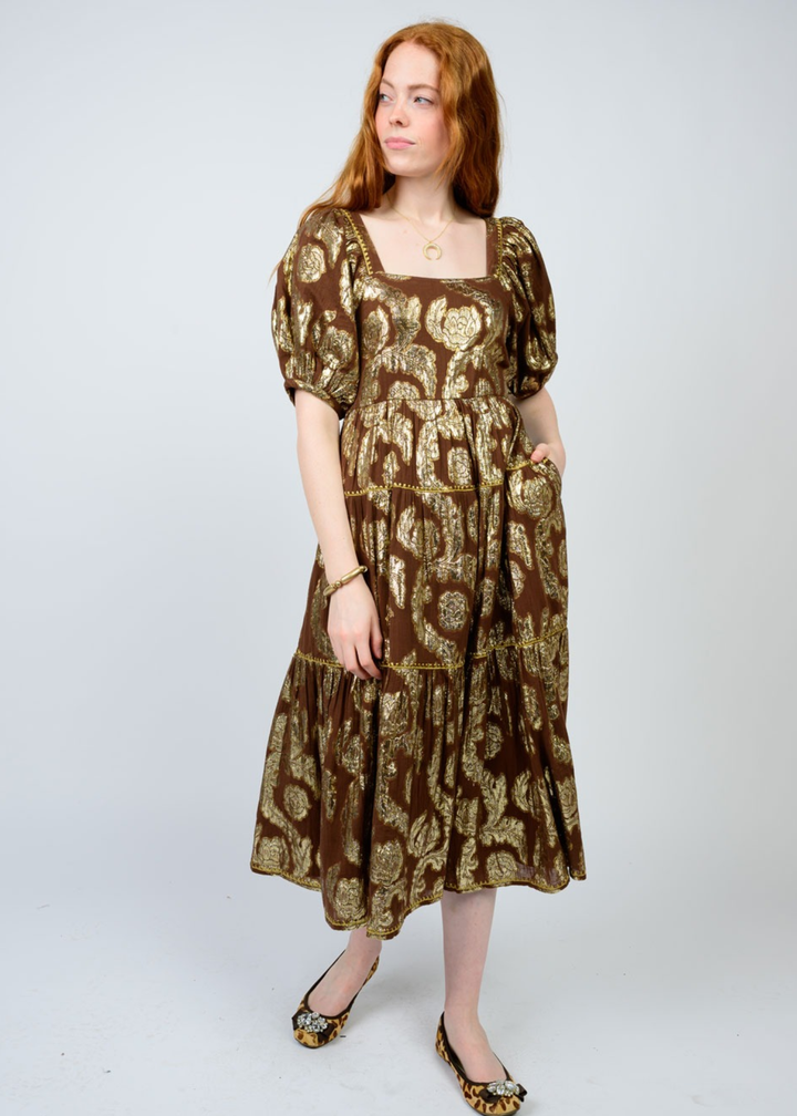Woman with hand in pocket wearing a brown midi dress with a gold floral pattern