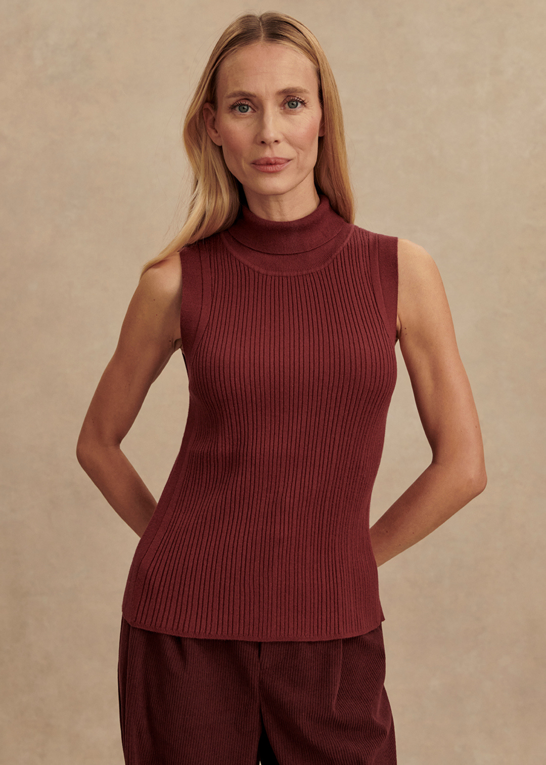 Woman wearing a maroon colored tank with brown pants in front of a tan background