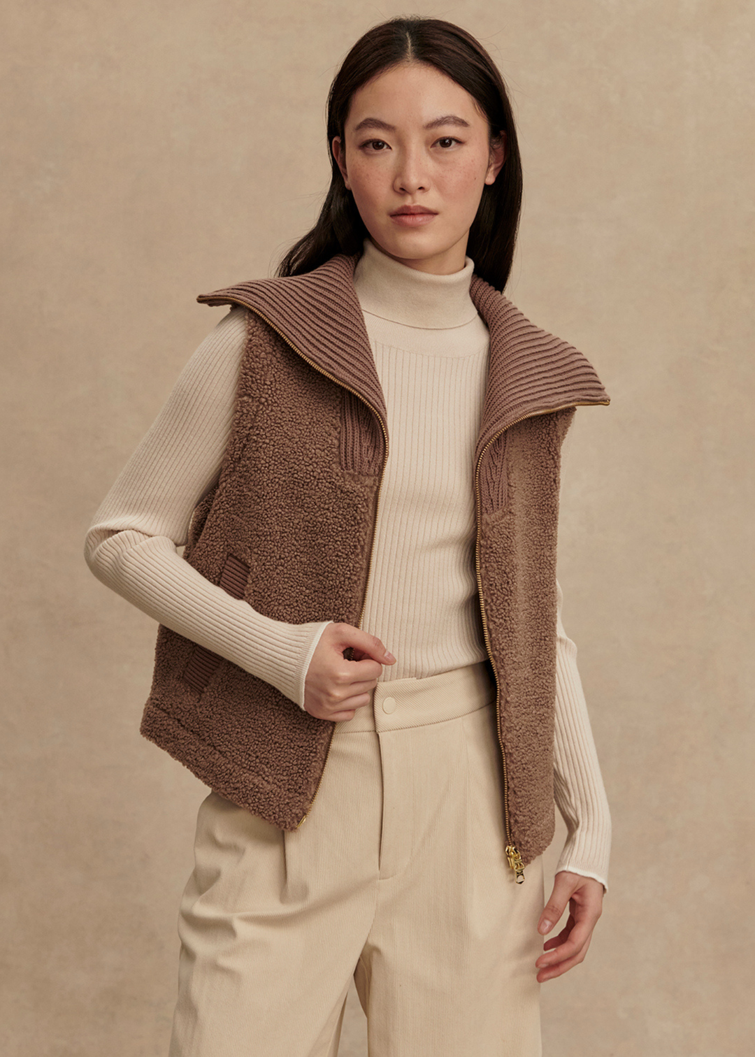 Woman wearing a brown vest over a cream turtleneck and cream pants standing in front of a tan background