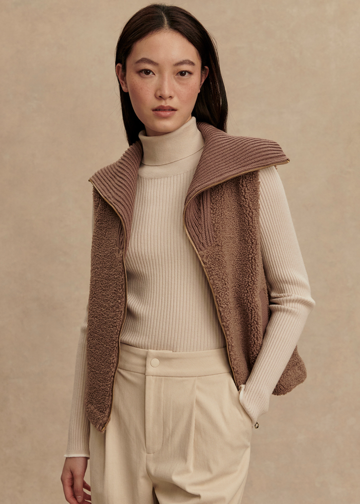 Woman wearing a brown vest over a cream turtleneck and cream pants with hand in pocket standing in front of a tan background