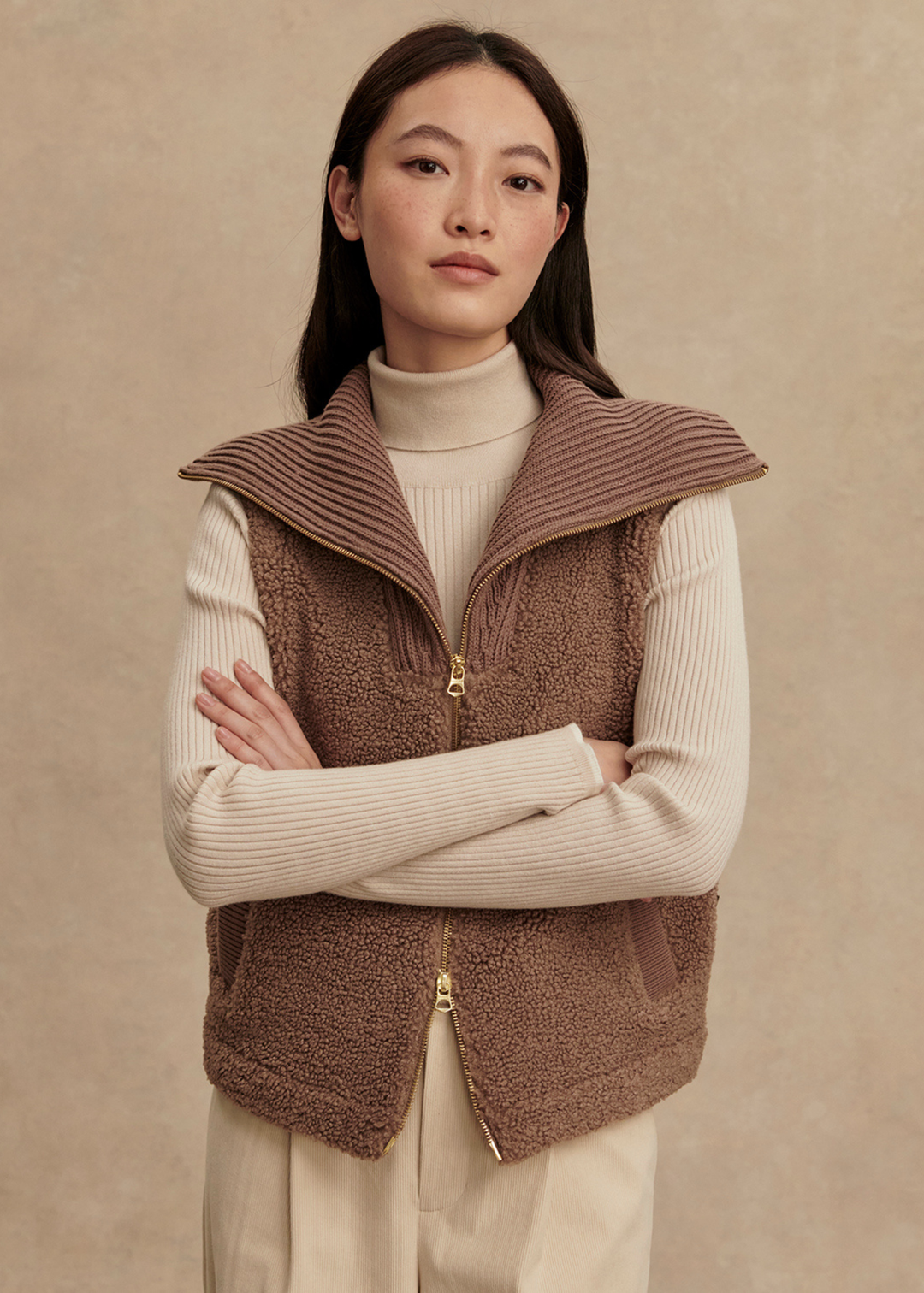 Woman with arms crossed wearing a brown vest over a cream turtleneck and cream pants standing in front of a tan background