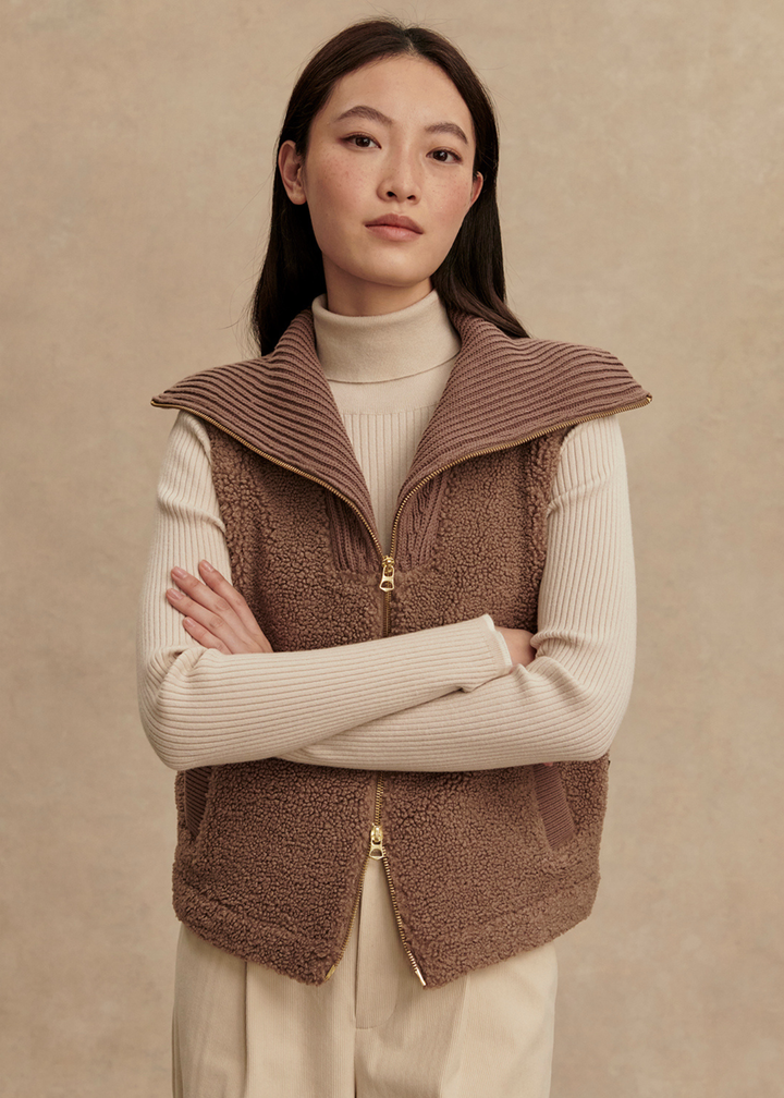 Woman with arms crossed wearing a brown vest over a cream turtleneck and cream pants standing in front of a tan background