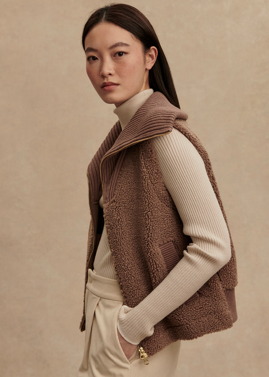 Woman turned to the side wearing a brown vest over a cream turtleneck and cream pants with hand in pocket standing in front of a tan background