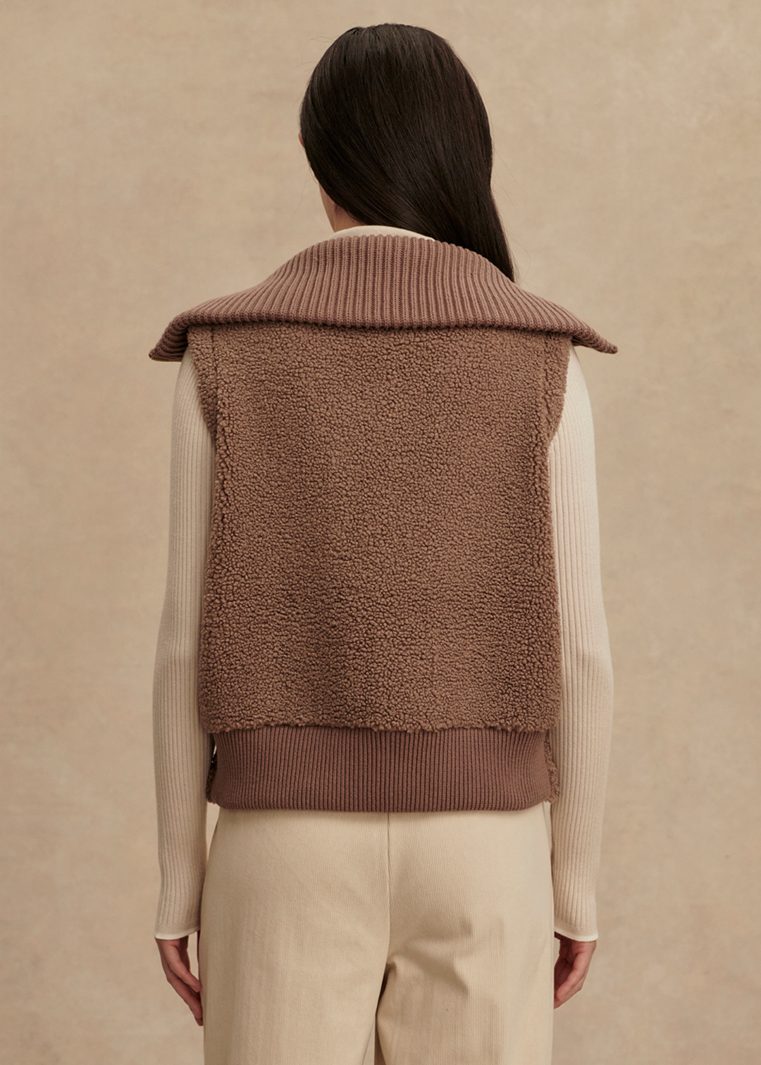Woman turned around wearing a brown vest over a cream turtleneck and cream pants standing in front of a tan background
