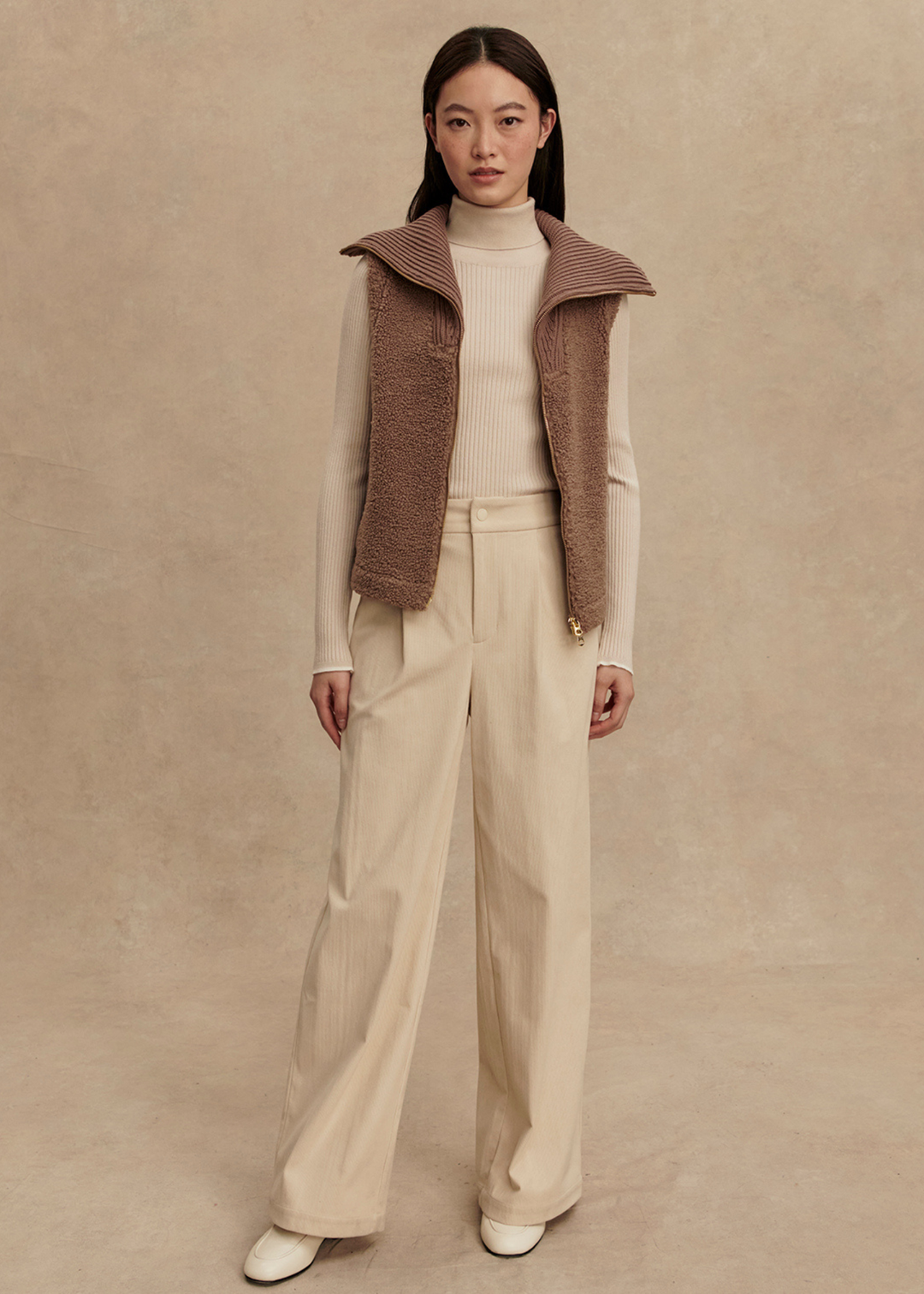 Woman wearing a brown vest over a cream turtleneck and cream pants standing in front of a tan background