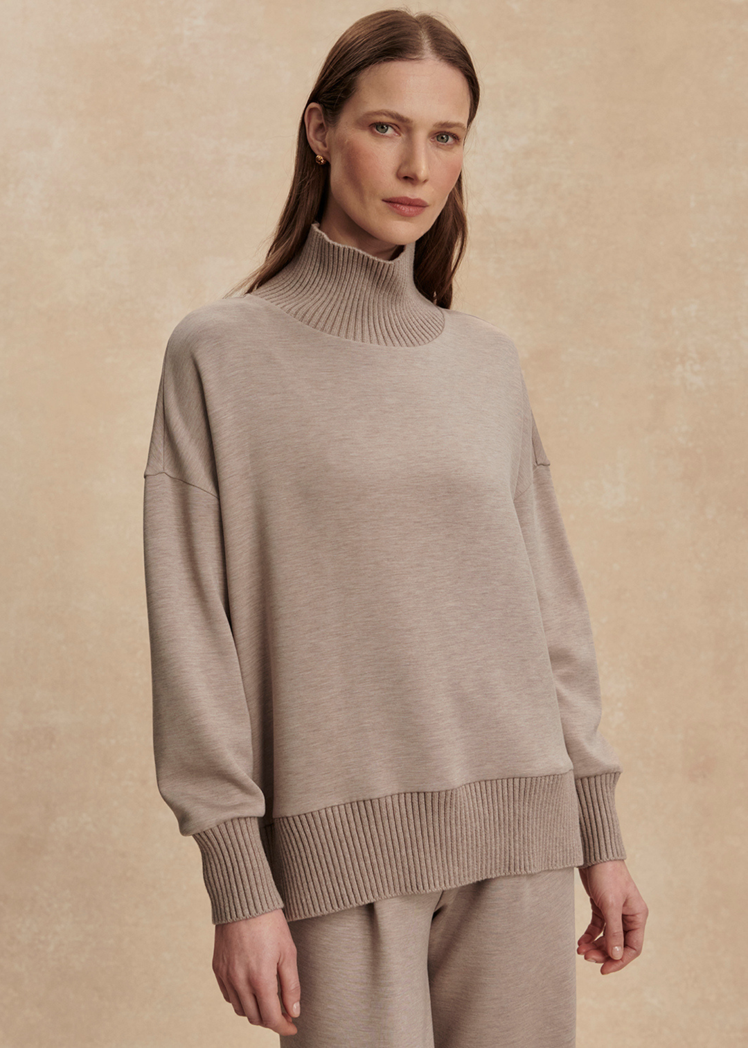 Woman wearing a taupe high neck sweatshirt with taupe pants in front of a tan background