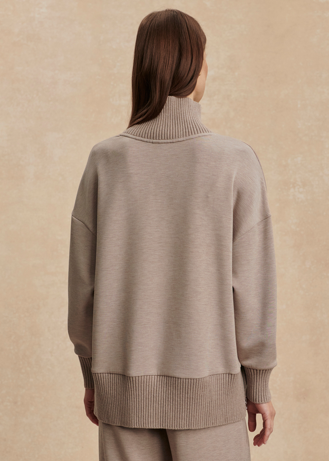 Woman turned around wearing a taupe high neck sweatshirt with taupe pants in front of a tan background
