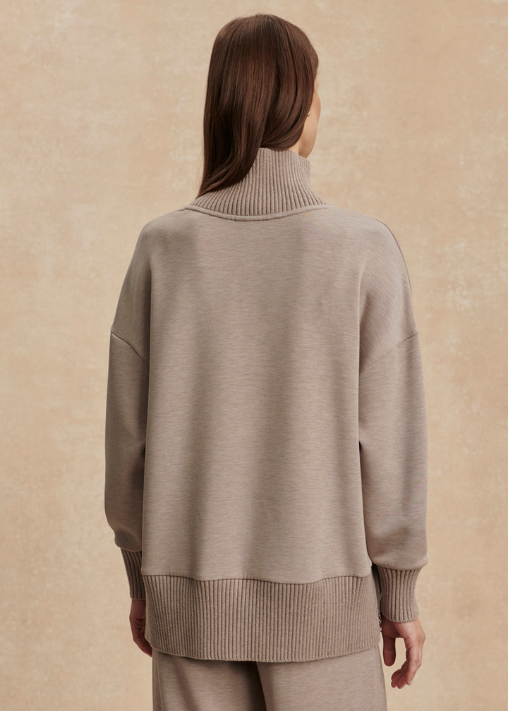 Woman turned around wearing a taupe high neck sweatshirt with taupe pants in front of a tan background