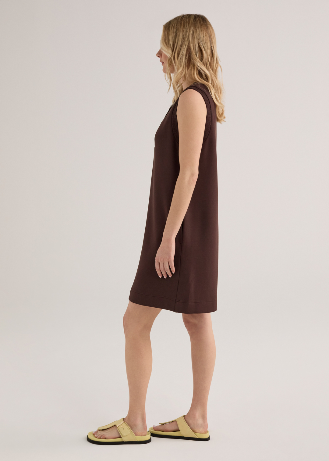 Woman turned to the side wearing a brown sleeveless mini dress with yellow sandals