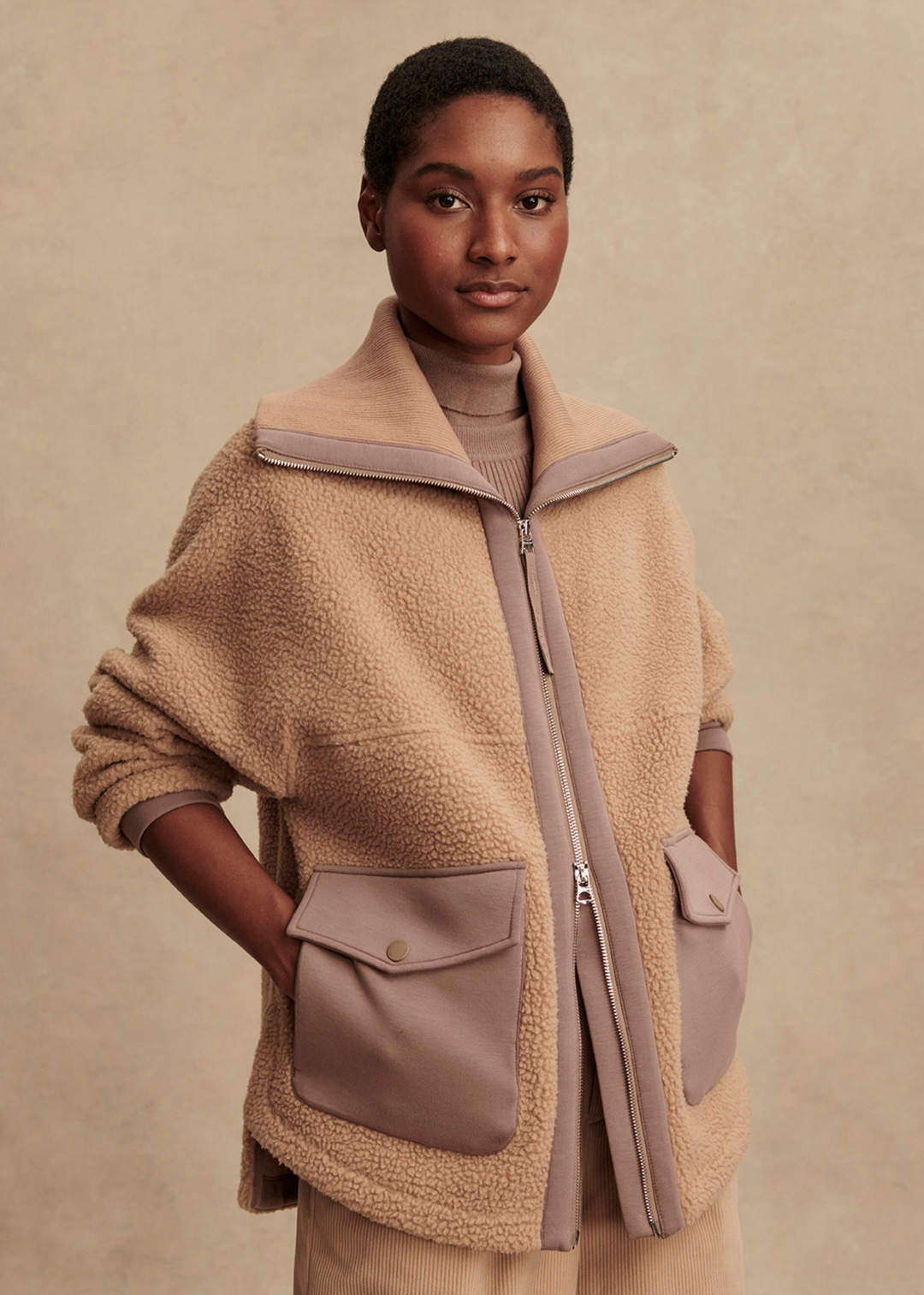 Woman wearing a tan zip up fleece jacket with taupe trim and pockets in front of a tan background