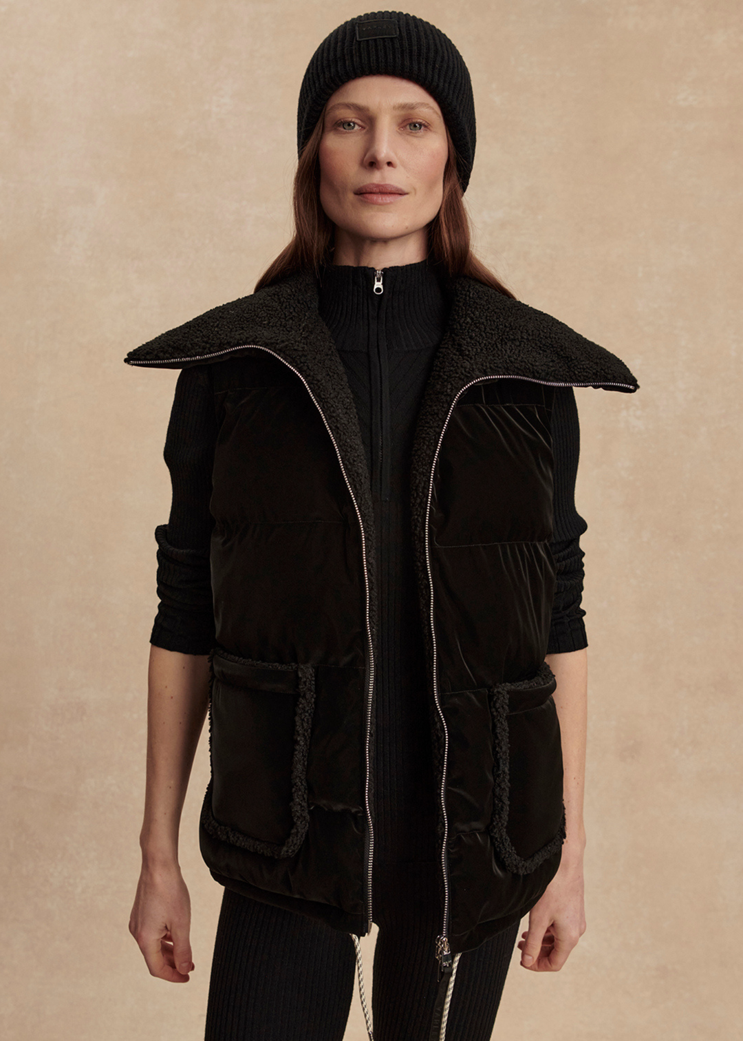 Woman wearing a black outfit with a black puffer vest against a tan background